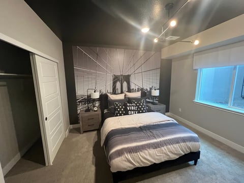 Modern bedroom with queen bed, white wardrobe, nightstands, Brooklyn Bridge wall art, gray carpet, and large windows