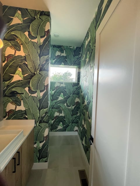 Bathroom hallway with green and gold tropical leaf wallpaper, white walls, vanity sink on left, and window at the end