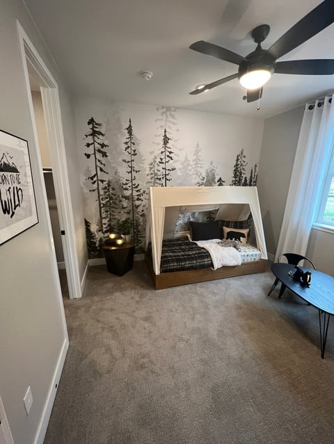 Modern bedroom with forest wall mural, wooden canopy bed, ceiling fan, desk, and minimalist décor in neutral tones