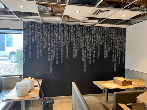 Modern restaurant interior with wooden tables, exposed ceiling, and large dark accent wall with white vertical text design