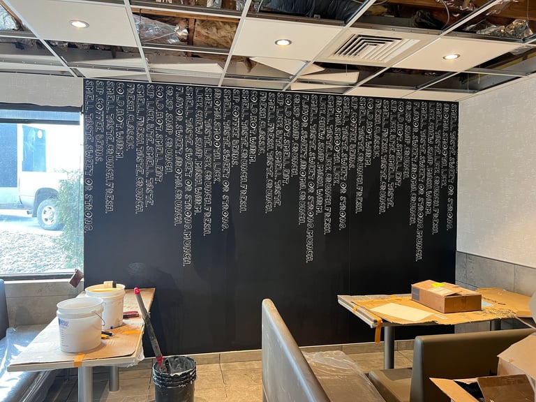 Modern restaurant interior with wooden tables, exposed ceiling, and large dark accent wall with white vertical text design