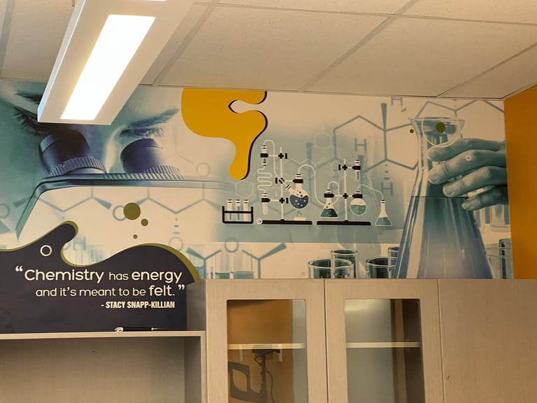 Science lab interior with blue hands mural, yellow signage, chemistry quote on wall, equipment, and white cabinetry below
