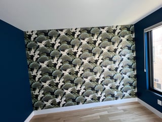 Modern bedroom interior with botanical wallpaper featuring white cranes and green foliage on accent wall, dark blue painted walls, and wooden flooring