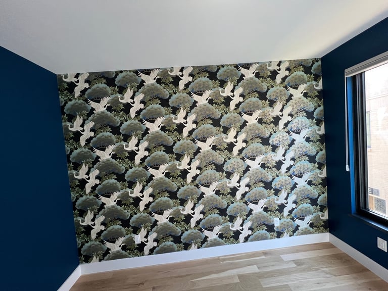 Modern bedroom interior with botanical wallpaper featuring white cranes and green foliage on accent wall, dark blue painted walls, and wooden flooring