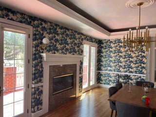 Modern dining room with navy floral wallpaper, fireplace, wood table, pendant lighting, and glass doors opening to an outdoor deck