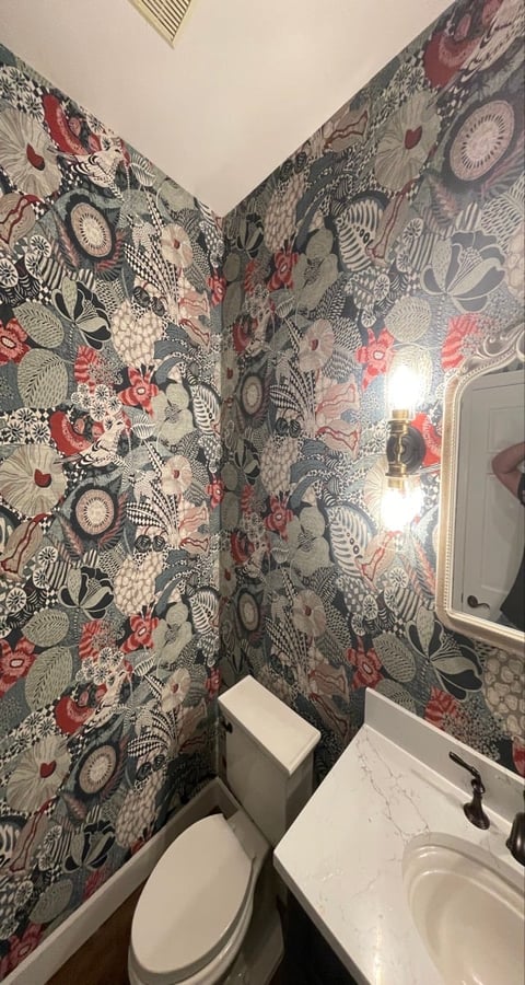 Small bathroom with patterned wallpaper featuring red flowers and botanical designs, white toilet, sink, and framed mirror