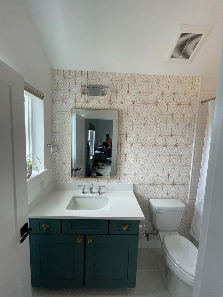 Modern bathroom with teal vanity, white countertop, rectangular mirror, geometric patterned wallpaper, toilet, and window
