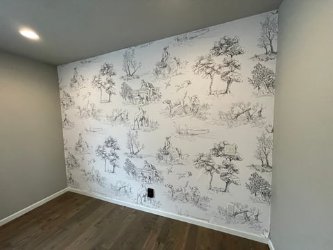 Interior room with one wall covered in toile de Jouy patterned wallpaper featuring sketched trees and pastoral scenes, hardwood flooring visible