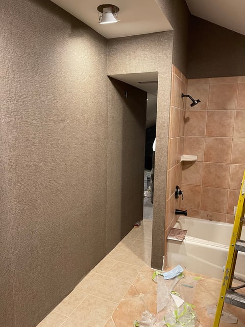 Bathroom undergoing renovation with tan textured walls, exposed tile work, bathtub visible on right, and construction materials on floor