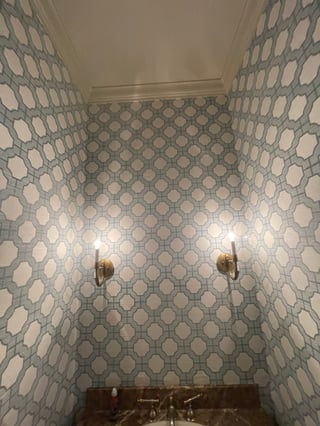 Small bathroom nook with blue and white geometric patterned wallpaper, two brass sconces, and dark vanity