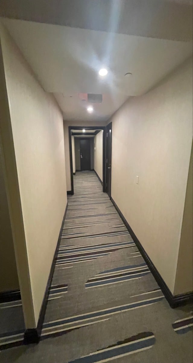 Long empty hallway with cream-colored walls, recessed ceiling lights, striped carpet flooring, and dark doorways along the sides