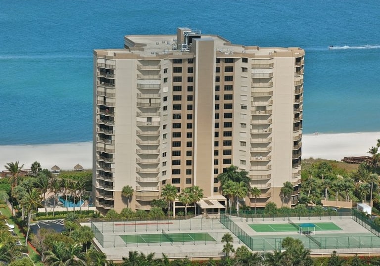 Beachfront high-rise residential building with tan and white exterior, palm trees, tennis courts, and turquoise ocean