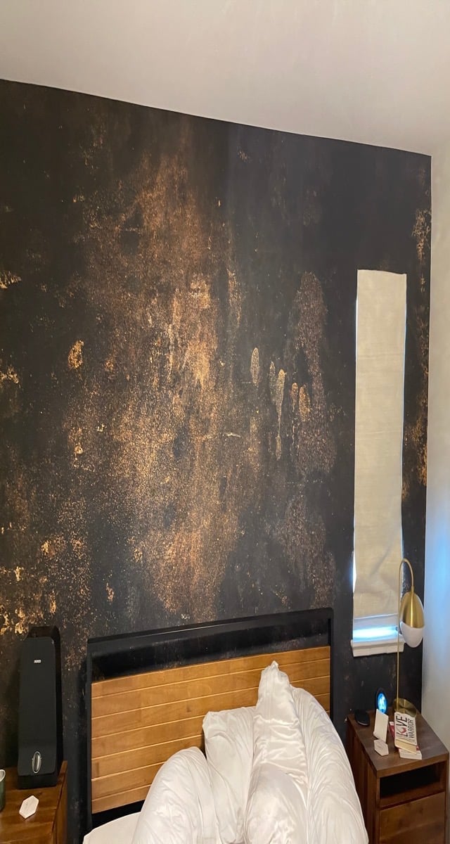 Bedroom with bed featuring wooden and black headboard against a large dark wall with distressed gold and rust patina texture