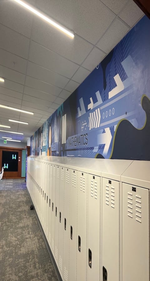 School hallway with white lockers beneath a large blue athletics mural featuring airplane graphics and fluorescent ceiling lights