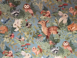 Woodland-themed fabric pattern featuring forest animals like deer, owls, raccoons, rabbits, and birds among green ferns and colorful flowers on a sage background