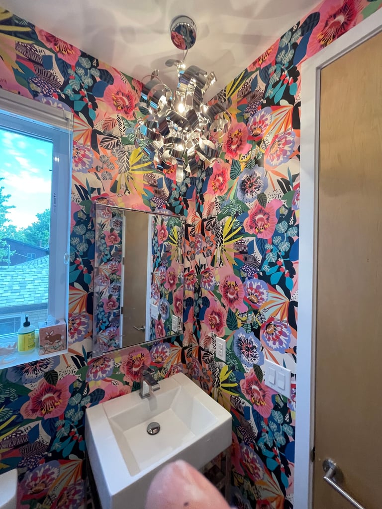 Colorful bathroom with vibrant pink and blue floral wallpaper, white pedestal sink, decorative crystal chandelier, and large mirror