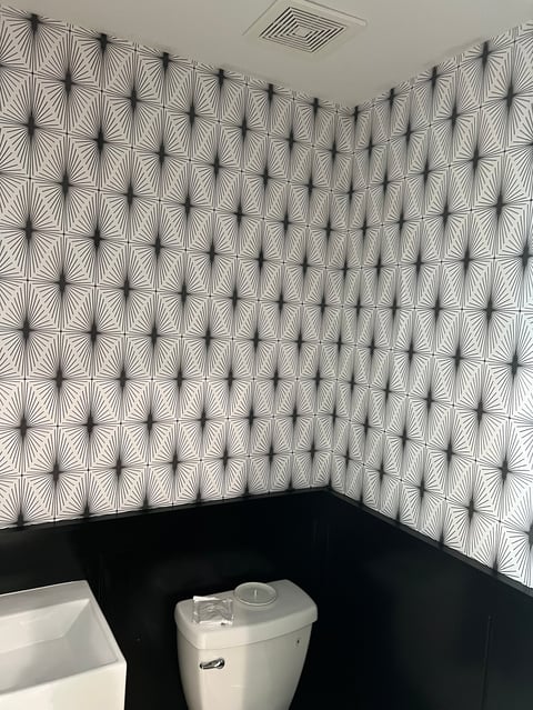 Bathroom corner with geometric patterned wallpaper featuring black starburst designs on white, white toilet, and ceiling vent
