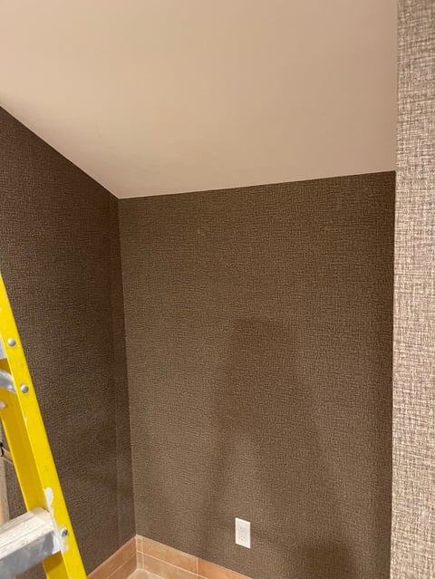 Interior corner of room with olive-brown textured wallpaper on two walls, cream ceiling, white outlet, and yellow ladder visible on left