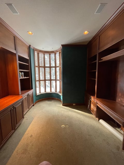 Empty office or study with curved bay window, built-in wooden cabinetry, shelving, and beige carpeted floor