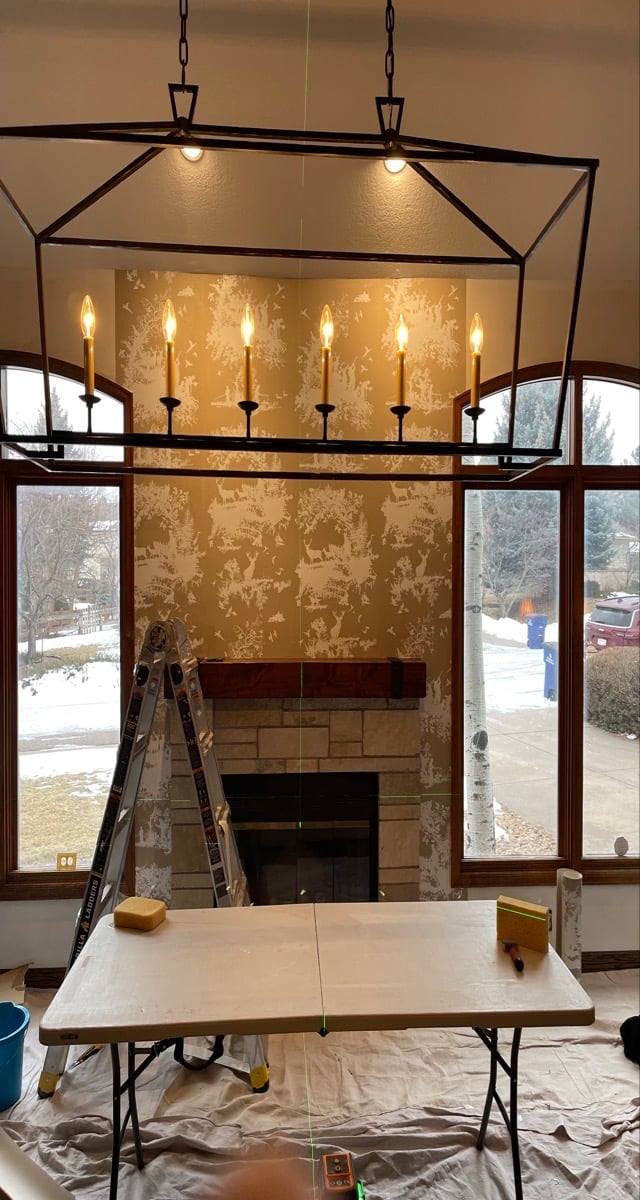 Modern lantern-style chandelier with six candle-style lights hanging above fireplace during renovation, with folding table in foreground