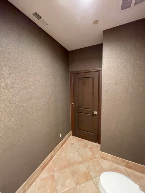 Small bathroom with brown textured walls, tile floor, dark wooden door, and white toilet in corner