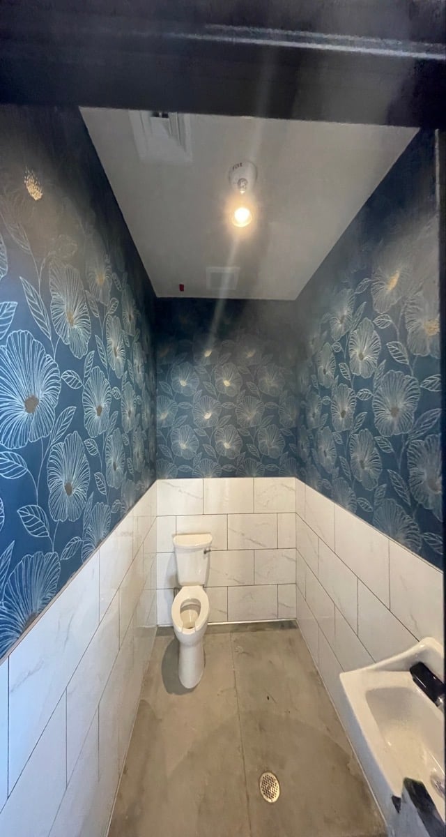 Narrow bathroom with white tile walls on lower half, dark blue floral wallpaper on upper half, toilet, recessed lighting, and polished concrete floor