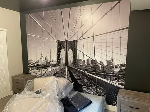 Black and white mural of Brooklyn Bridge spanning entire bedroom wall above unmade bed with plastic sheets