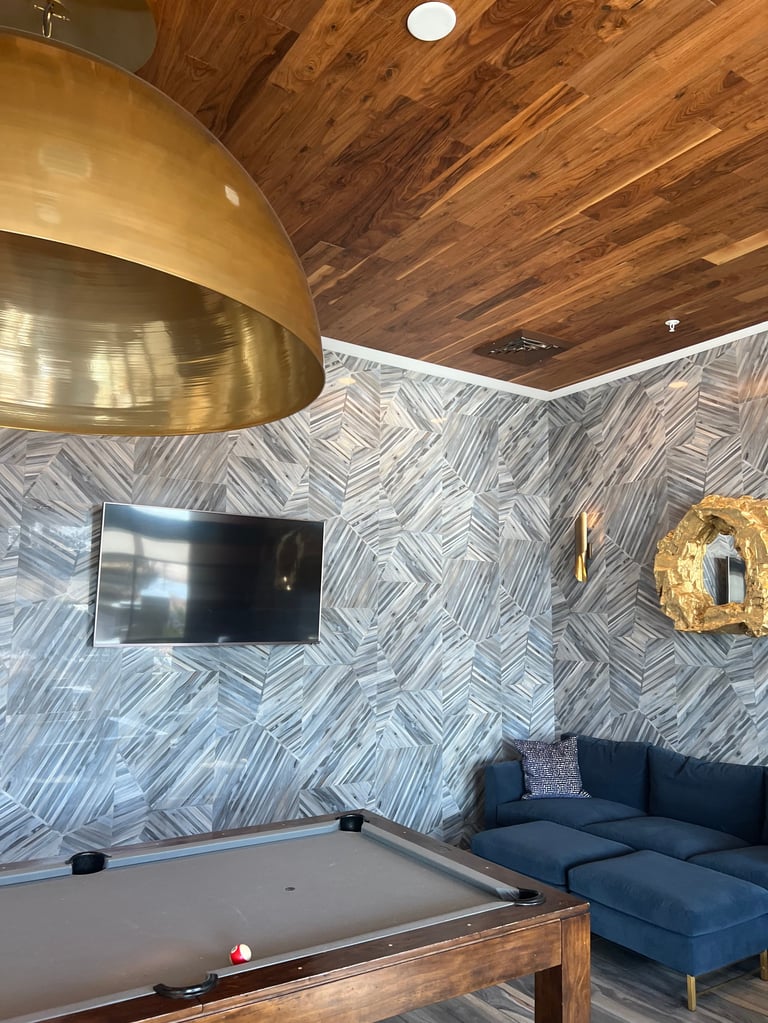 Modern game room with pool table, blue sectional sofa, mounted TV, geometric patterned walls, wood ceiling, and brass pendant lamp