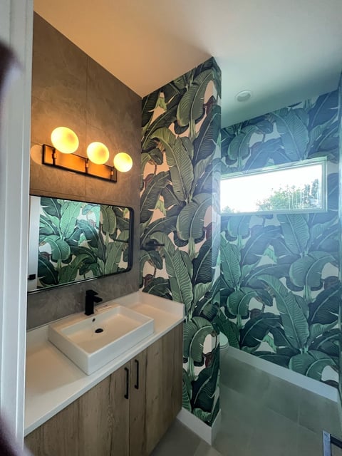 Modern bathroom with tropical wallpaper, white sink, wooden vanity, illuminated mirror, and bathtub with window view