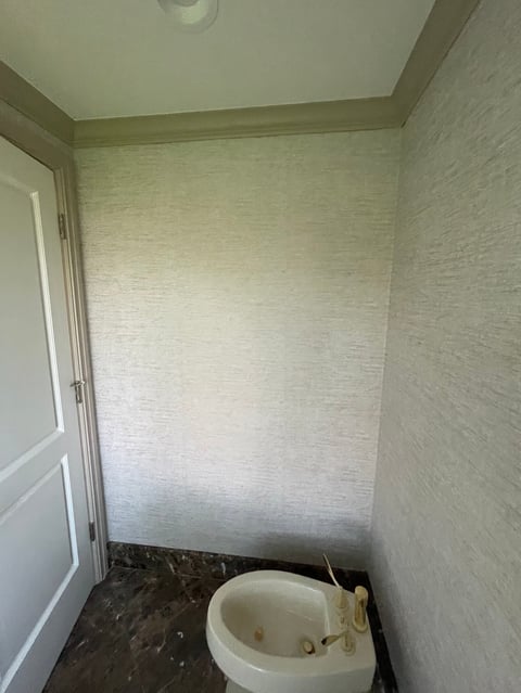 Partially renovated bathroom with white textured wallpaper, cream ceiling trim, and a white toilet on dark flooring in corner of room